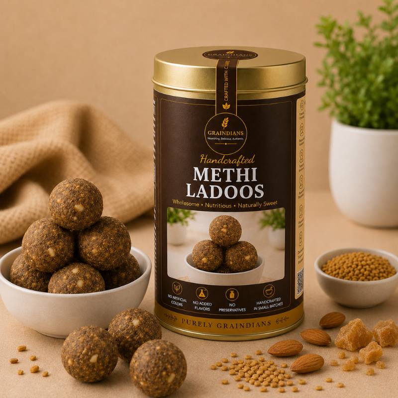 Graindians Homemade Methi Ladoos in a premium gift tin with fresh fenugreek ladoos, methi seeds, almonds and jaggery displayed on a warm beige background.